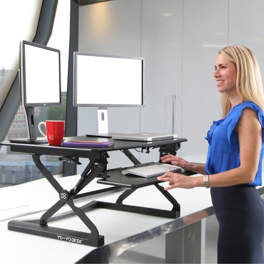 A black manual desk riser with a worktop above and a keyboard tray below, sitting on a desk.